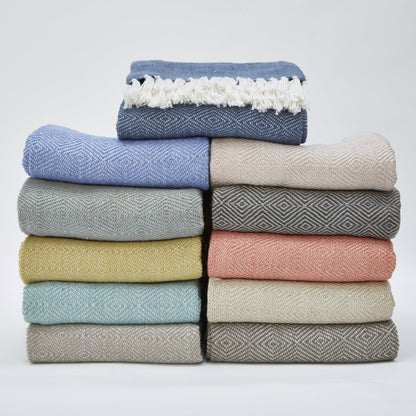 A neatly stacked pile of twelve woven blankets in assorted colours and patterns, topped with a blue fringed, machine-washable throw.