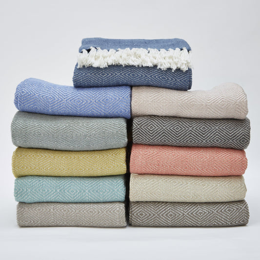 A neatly stacked pile of twelve woven blankets in assorted colours and patterns, topped with a blue fringed, machine-washable throw.
