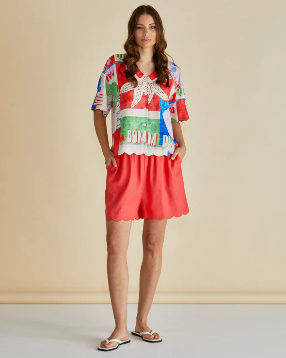 Woman standing against a plain background, wearing a colourful seashell print shirt with patterns and red scalloped shorts, hands in pockets, and white sandals made from vegan materials.