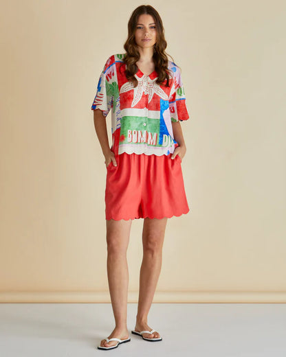 Woman standing against a plain background, wearing a colourful seashell print shirt with patterns and red scalloped shorts, hands in pockets, and white sandals made from vegan materials.