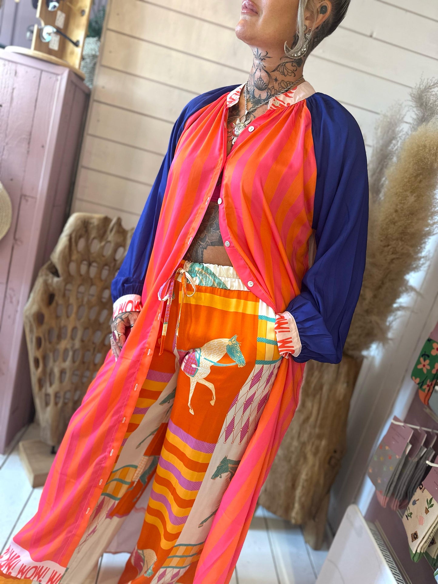A person wearing a bright outfit with a pink and orange striped top, blue sleeves, and patterned trousers featuring horses, standing indoors with decorative elements in the background.