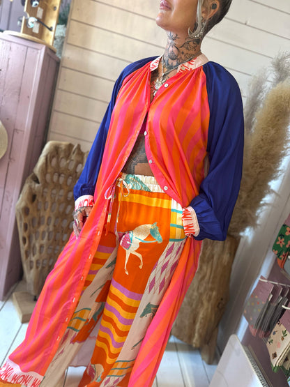 A person wearing a bright outfit with a pink and orange striped top, blue sleeves, and patterned trousers featuring horses, standing indoors with decorative elements in the background.