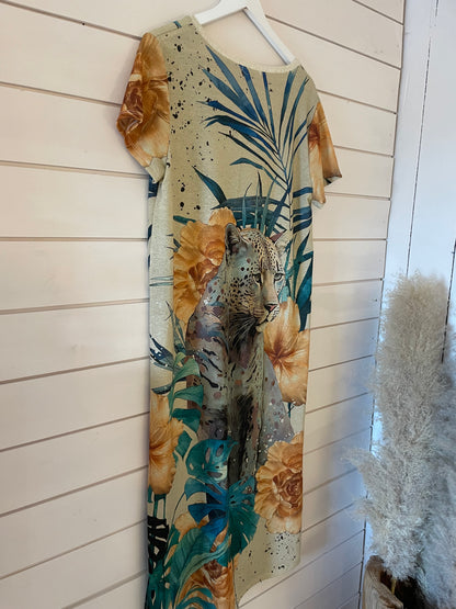 A short-sleeved evening dress featuring a leopard, tropical leaves, and large orange flowers printed on shimmering fabric hangs against a white wooden wall.