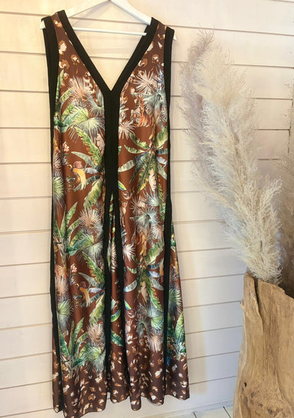 Sleeveless brown Jungle Jumpsuit Dress with a tropical leaf and feather print hangs on a white hanger against a shiplap wall, beside pampas grass in a wooden vase. Boasts versatile fit and breathable fabric.