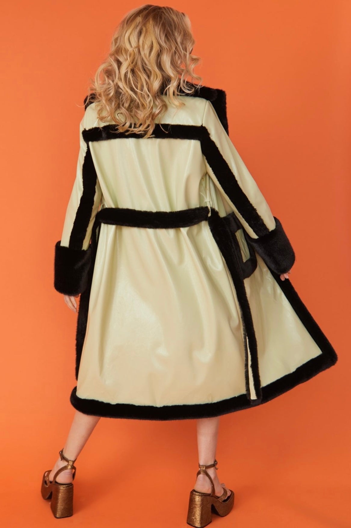 A person with wavy blonde hair models a light-coloured belted coat featuring black trim and leopard print lining, standing against an orange background.