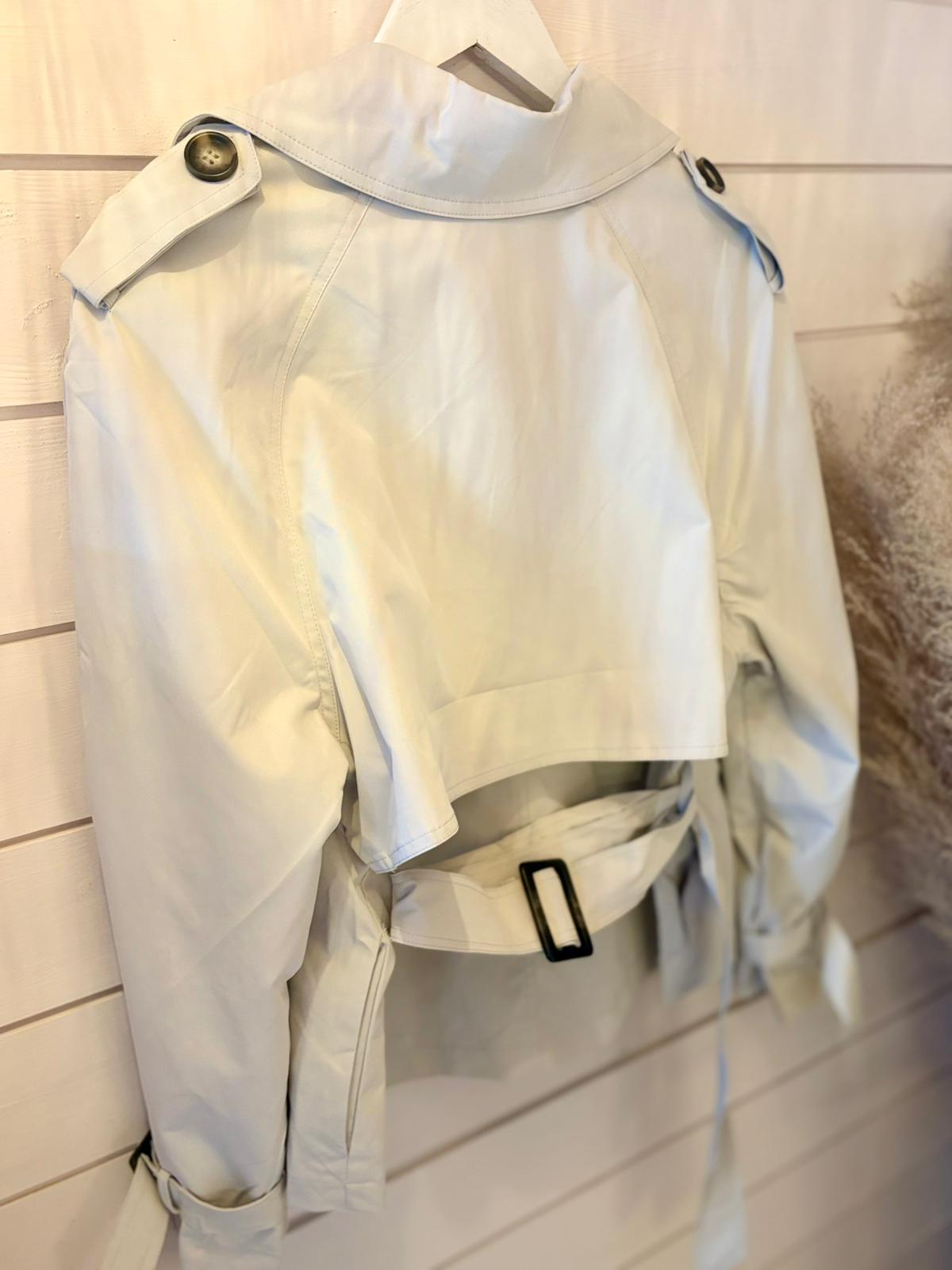 A light beige cropped trench coat with a belt and shoulder epaulettes hangs on a white wall-mounted hanger.