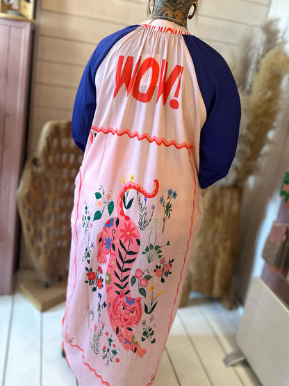 Person wearing a long pink and blue dress with WOW! in red on the back and a large floral pink tiger illustration at the hem, standing indoors.