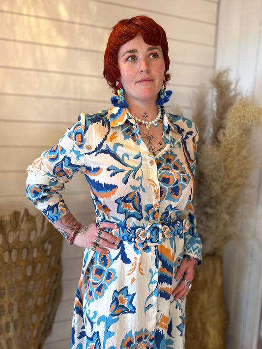 A person with short red hair, wearing a cream shirt dress with long sleeves and blue earrings, stands indoors near a decorative basket and dried pampas grass—perfect for breezy summer days.