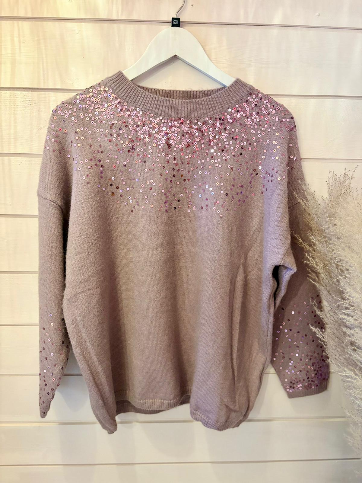 A purple sequin jumper crafted from vegan materials, adorned with pink and silver sequins scattered over the shoulders and sleeves, hangs on a white hanger against a wooden wall.
