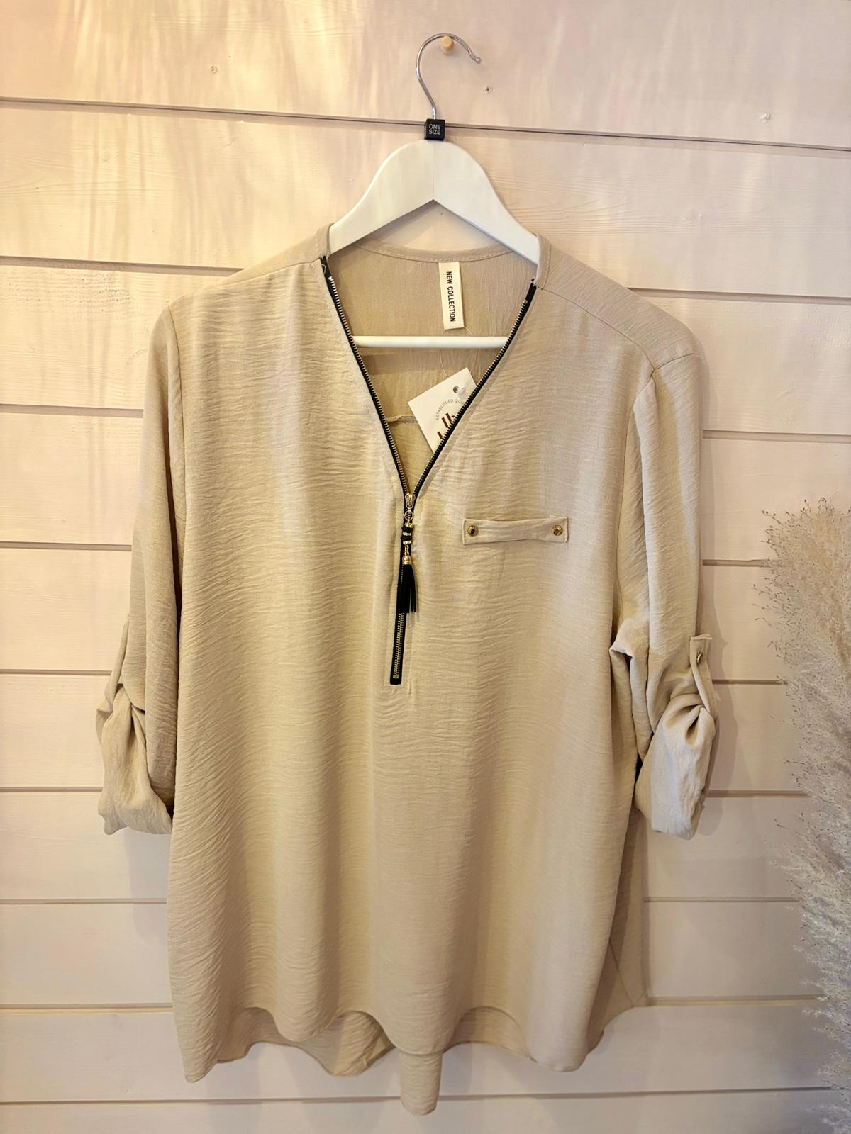 Beige long-sleeved blouse featuring a front zip detail and chest pocket, shown on a white hanger against a wooden wall.
