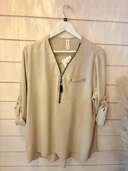 Beige long-sleeved blouse featuring a front zip detail and chest pocket, shown on a white hanger against a wooden wall.