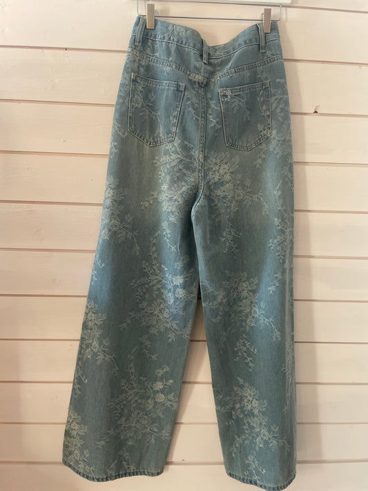 Super-soft blue floral jeans with a subtle flower pattern; these wide-leg jeans are displayed from the back, hanging on a wooden wall.
