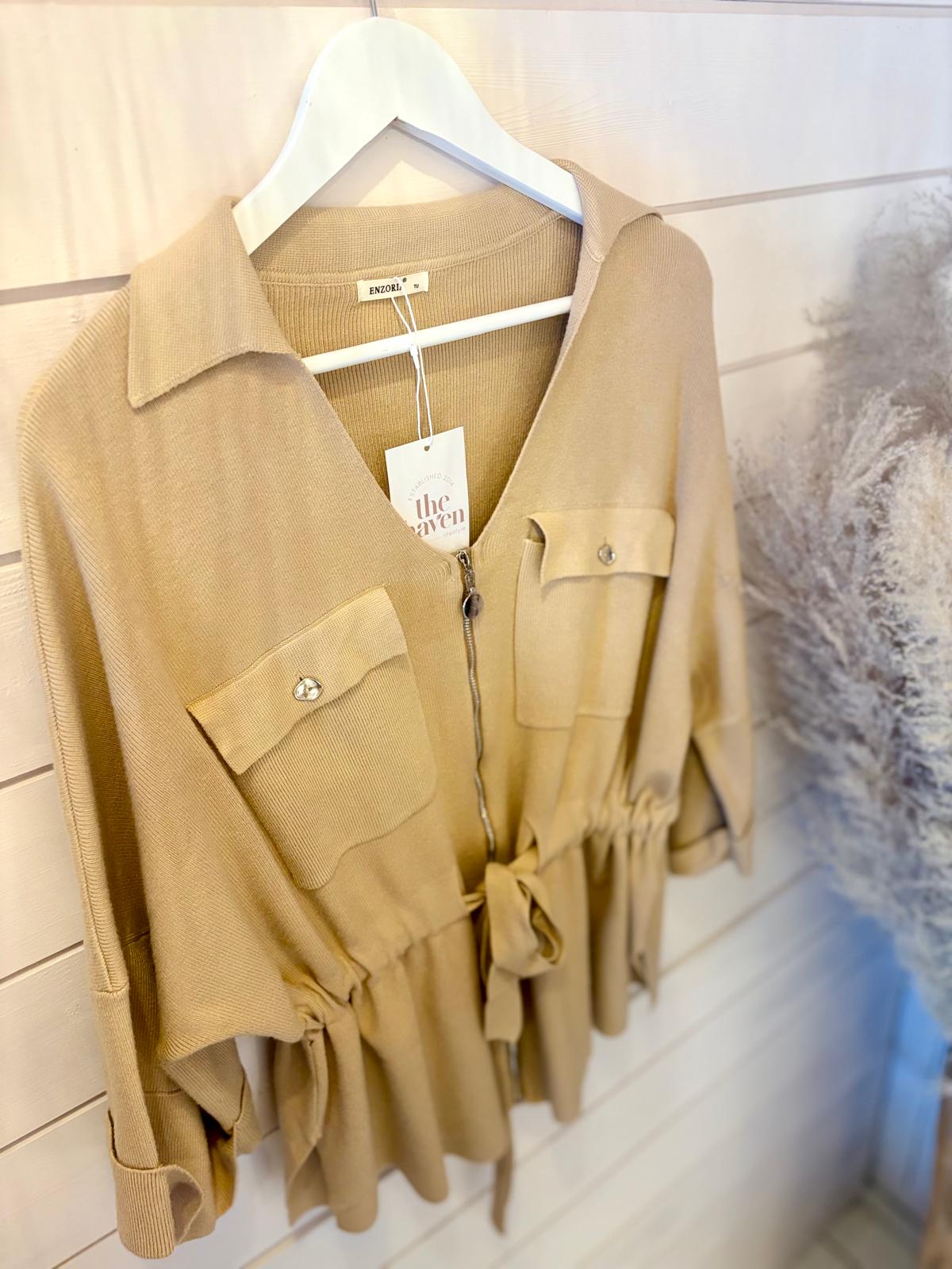 A beige long-sleeved button-up shirt with two front pockets and a tie waist, hanging on a white hanger against a wooden wall.