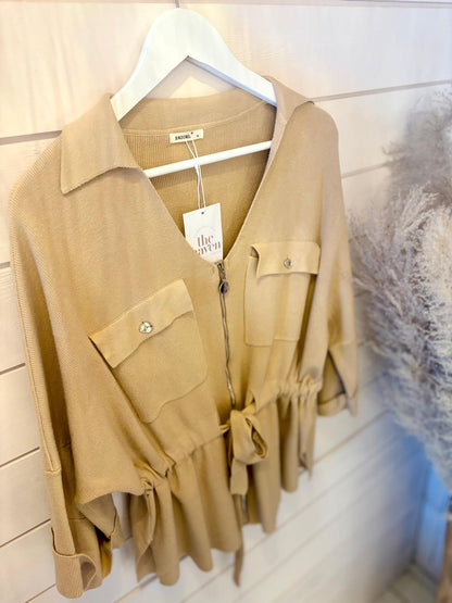 A beige long-sleeved button-up shirt with two front pockets and a tie waist, hanging on a white hanger against a wooden wall.