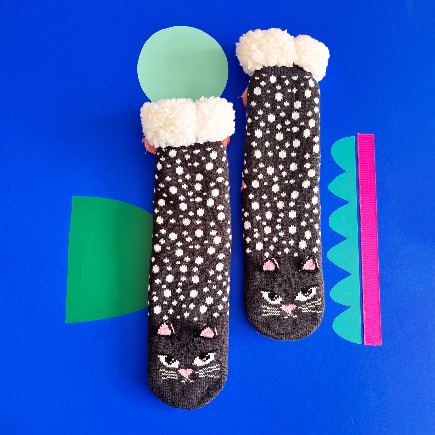 A pair of Cat Face Slipper Socks with white spots and fluffy cuffs is laid out on a blue background with geometric paper shapes—perfect as a cosy gift for cat lovers.