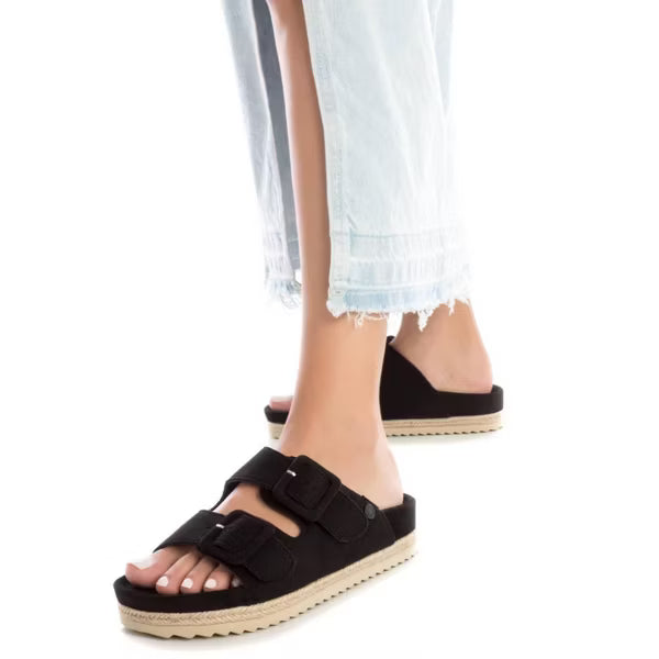 Person wearing frayed light-wash jeans and black vegan suede sandals with double straps and a flatform sole, standing against a white background.