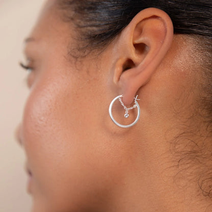 Close-up of a person’s ear adorned with silver hoop earrings featuring a small dangling gemstone and decorative detailing.