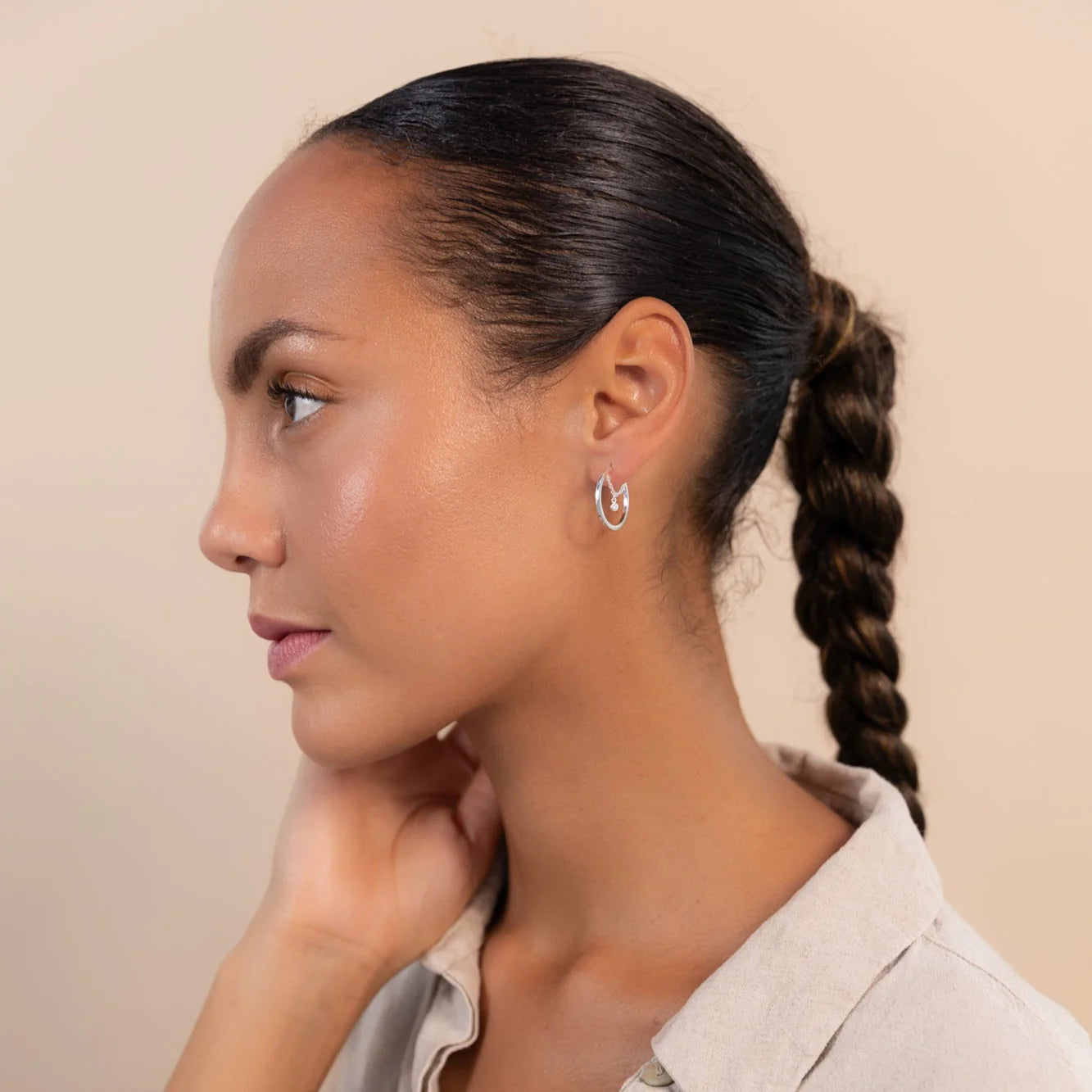 A woman with a tidy braided ponytail wears silver hoop earrings and a beige collared shirt, gazing to the left against a plain background.
