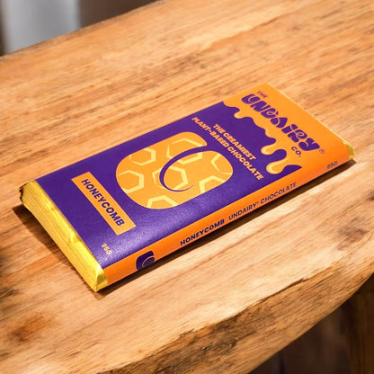 A chocolate bar labelled Honeycomb in purple and yellow packaging rests on a wooden surface. This ethically conscious treat is plant-based, vegan, and non-dairy chocolate, weighing 85 grams.