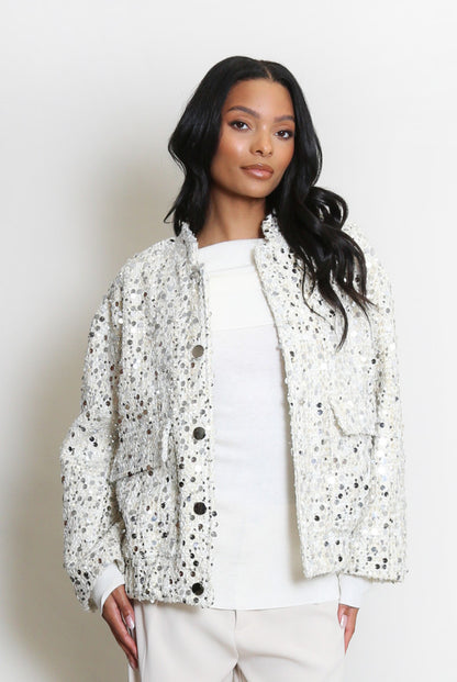 A woman stands against a plain backdrop wearing a vegan-friendly, white sequin jacket over a white top and light-coloured trousers.