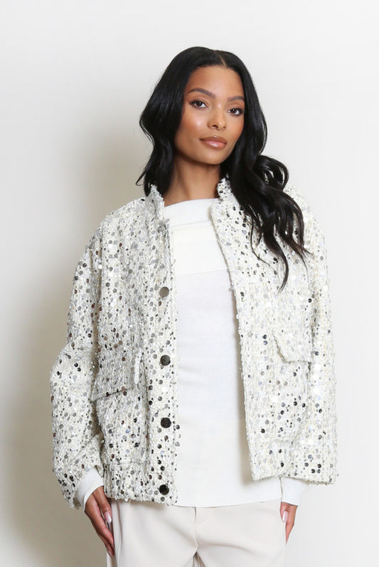 A woman stands against a plain backdrop wearing a vegan-friendly, white sequin jacket over a white top and light-coloured trousers.