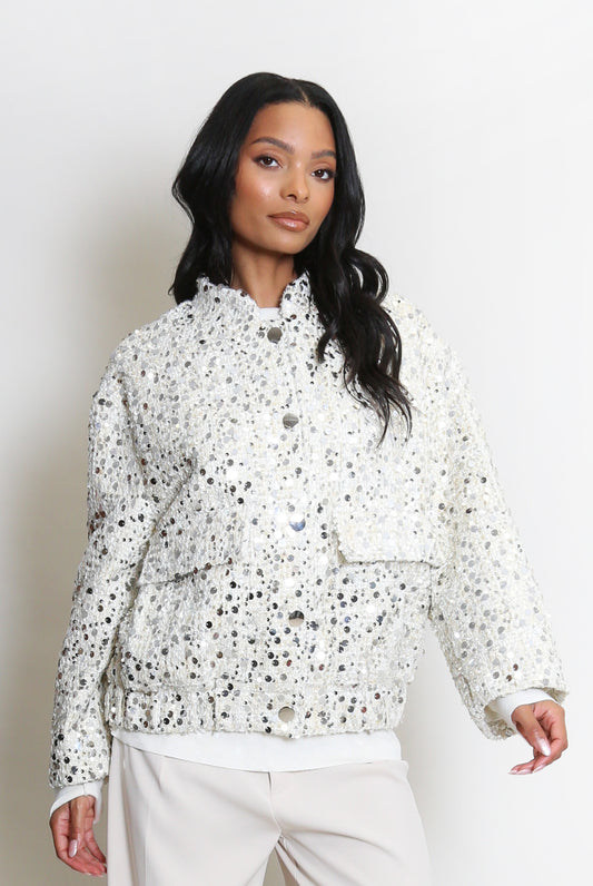 A woman dressed in a white sequin jacket with silver embellishments and large front buttons, teamed with cream trousers, stands against a plain white background.