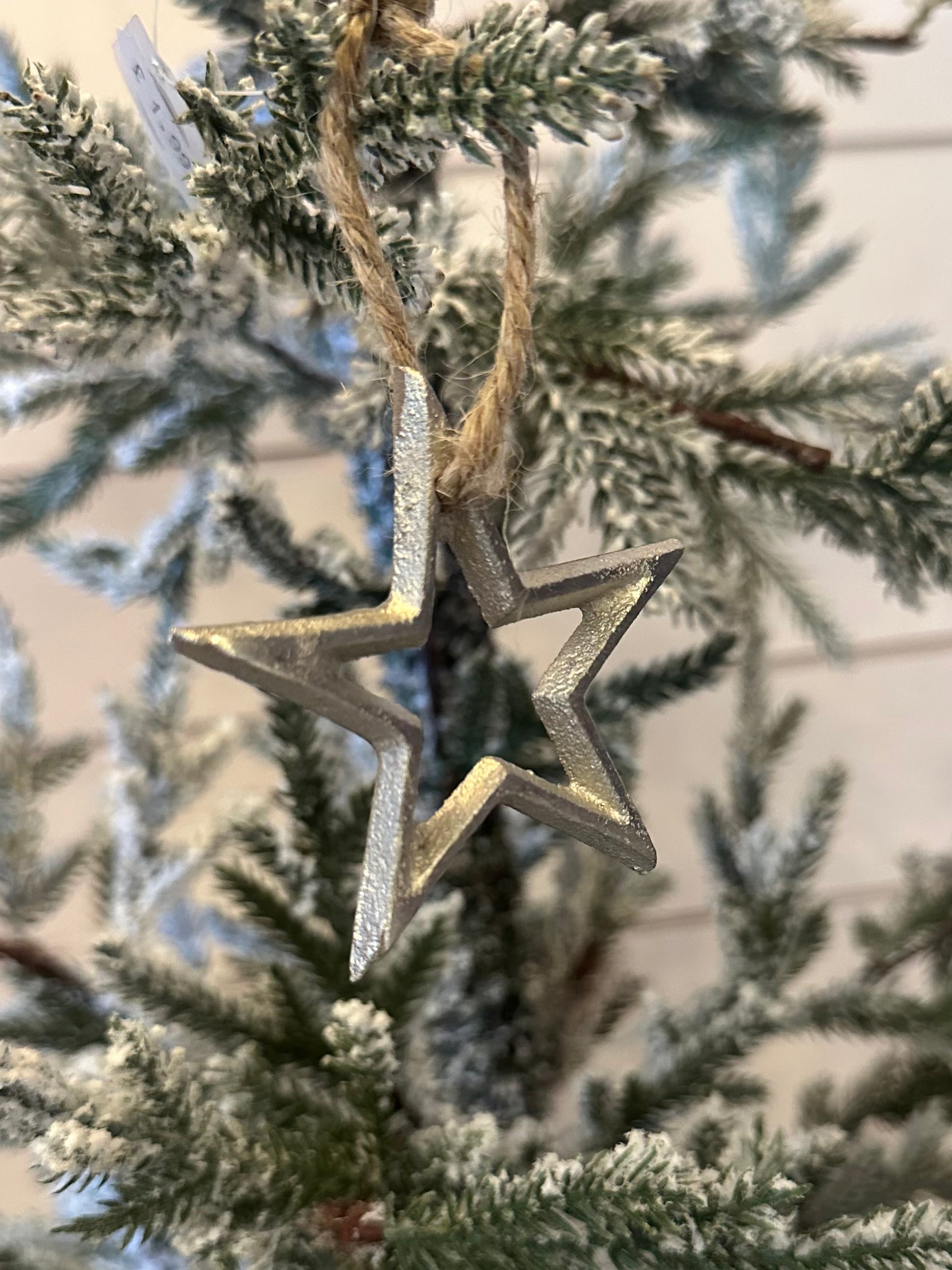 A silver Christmas star tree decoration hangs from a twine loop on a snow-dusted artificial tree, ideal for those looking for environmentally friendly Christmas décor.