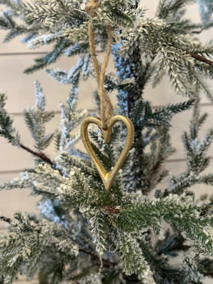 A gold, vegan-friendly Christmas heart tree decoration hangs by a twine string on a snow-dusted artificial evergreen, adding festive charm to your holiday décor.