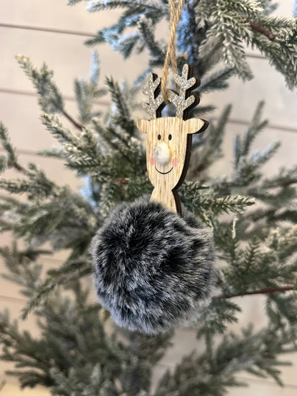 A charming deer decoration with glittery antlers and a large, fluffy, grey pom-pom body hangs on a snow-dusted artificial Christmas tree, adding festive holiday décor to your space.