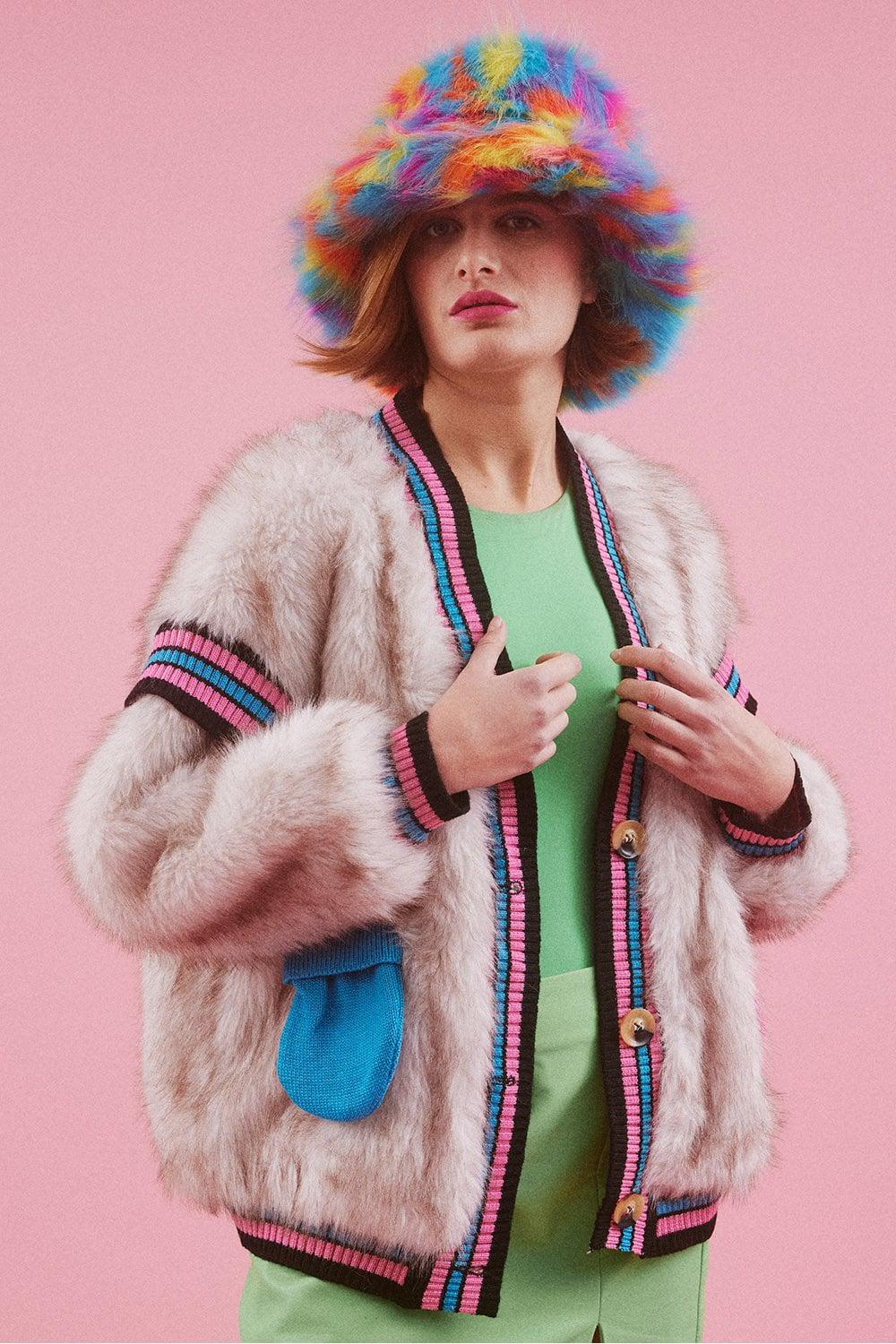 A person wearing a furry beige jacket with pink and blue trim, a green top, and a rainbow-coloured bucket hat made from cruelty-free vegan materials stands against a pink background.