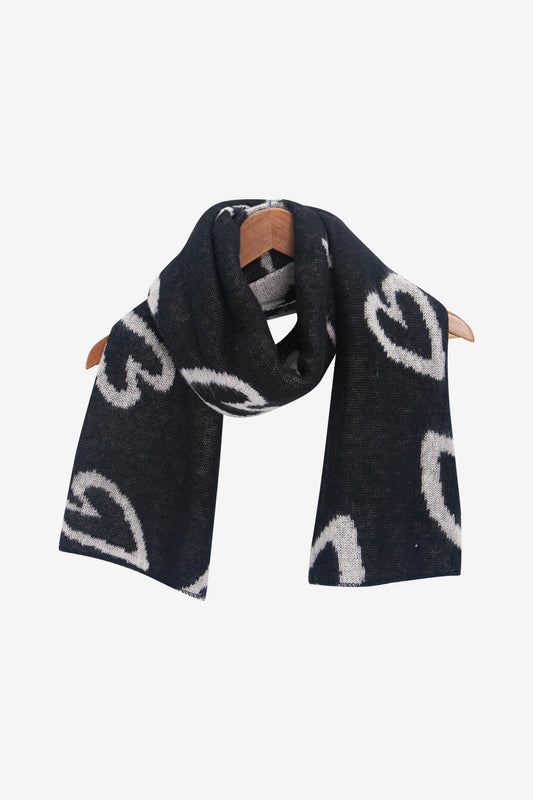 A vegan heart scarf, made of black fabric decorated with a white heart pattern, is elegantly draped over a wooden hanger set against a plain background.