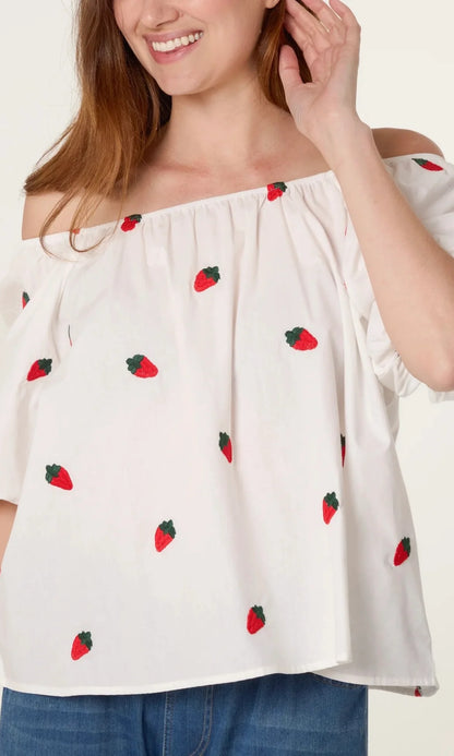 A woman in a Strawberry Ivory Puff Sleeve Top adorned with red strawberry prints and blue jeans, smiling as she touches her hair.