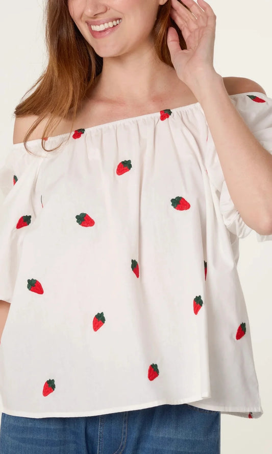 A woman in a Strawberry Ivory Puff Sleeve Top adorned with red strawberry prints and blue jeans, smiling as she touches her hair.