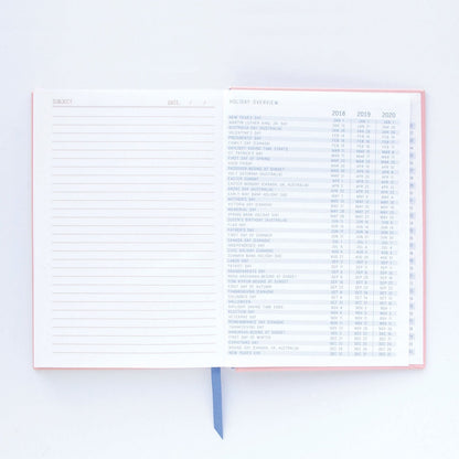 An open premium notebook with lay-flat binding, displaying a lined notes page on the left and a holiday overview for 2018 to 2020 on the right, along with a blue ribbon bookmark.