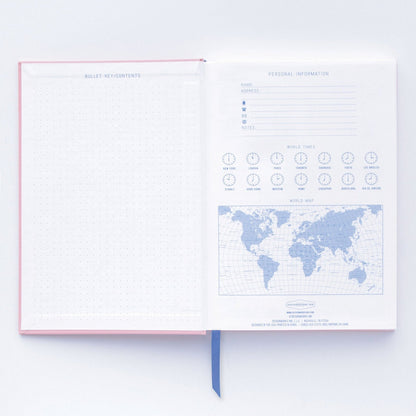 An open premium notebook with a soft-touch cover shows a dotted grid page to the left and, on the right, a personal information form, world clocks, and a world map.