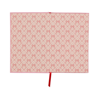 An open lay-flat journal with pink covers and pages displaying a repeating pattern of red bows, along with a red ribbon bookmark positioned in the centre.