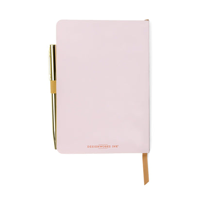 A closed pale pink gratitude journal with DESIGNWORKS INK on the cover, featuring an elastic pen loop holding a gold pen and a tan ribbon bookmark—ideal for your mindfulness or wellbeing routine.