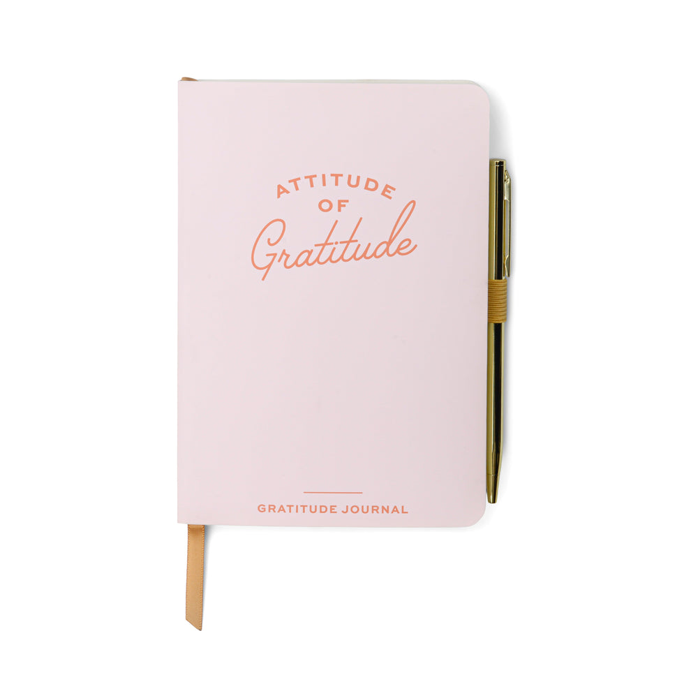 A closed pink gratitude journal with Attitude of Gratitude on the cover, a gold pen clipped to the side, and a beige ribbon bookmark—ideal for enhancing mindfulness in your daily wellbeing routine.