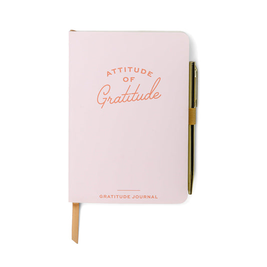 A closed pink gratitude journal with Attitude of Gratitude on the cover, a gold pen clipped to the side, and a beige ribbon bookmark—ideal for enhancing mindfulness in your daily wellbeing routine.