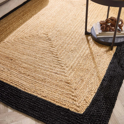 A woven jute rug with a black border rests on a wooden floor beside a round side table holding books and decorative items, bringing sustainable style to the room.