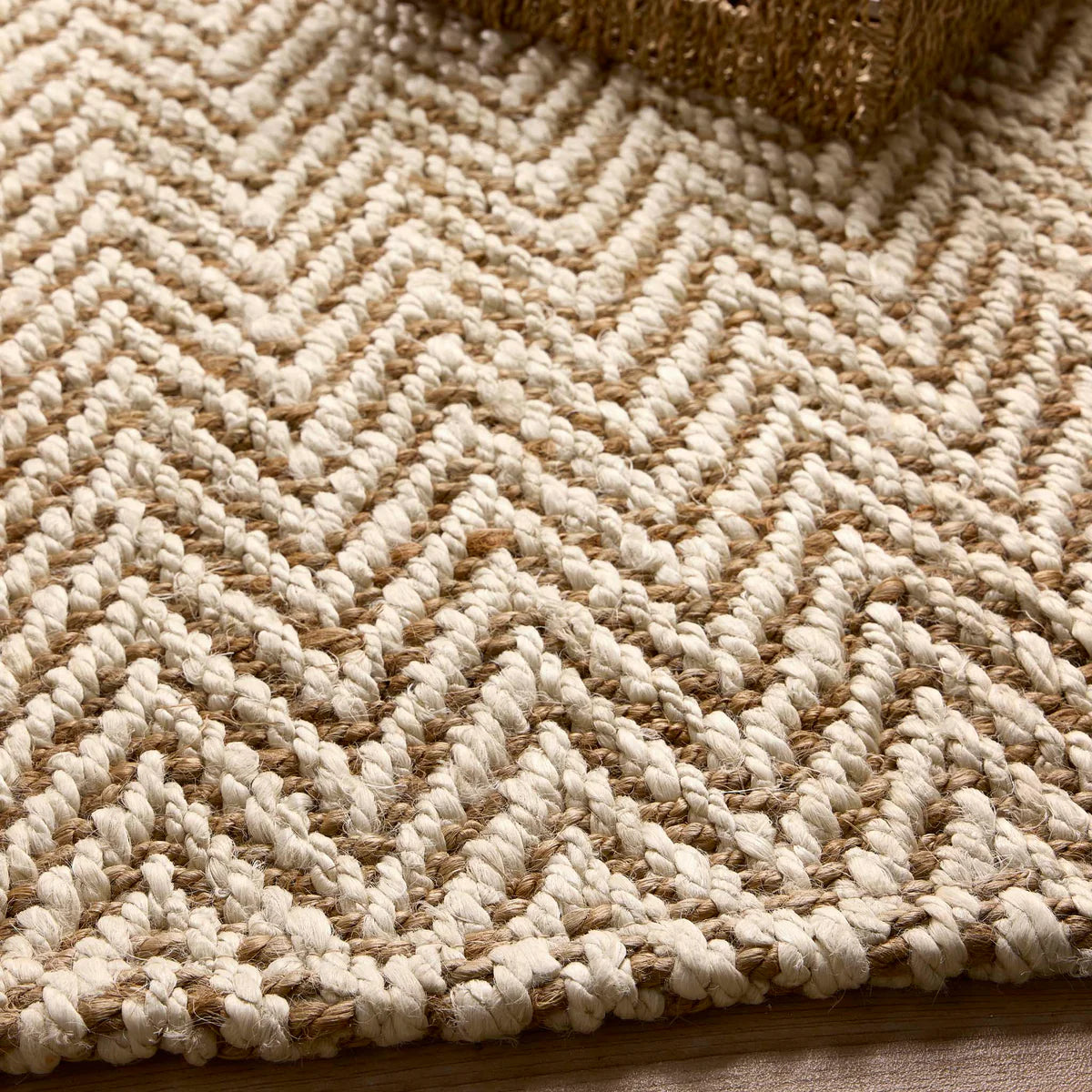 Close-up of a handmade rug featuring a zigzag chevron pattern in beige and tan; a textured object is visible in the top right corner.