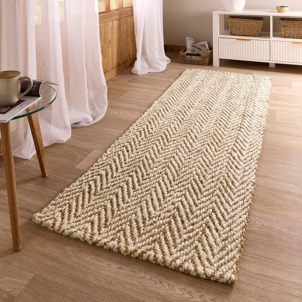 A natural jute rug with a herringbone weave rests on a light wooden floor in a bright, cosy room with sheer curtains and wicker baskets atop a white cabinet.
