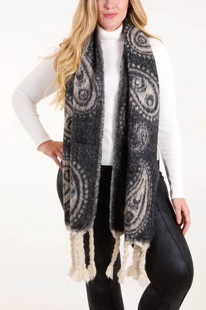 A woman in a white polo neck and black trousers poses with a vegan black paisley scarf adorned with braided tassels and an elegant paisley design, made from cruelty-free fabric.
