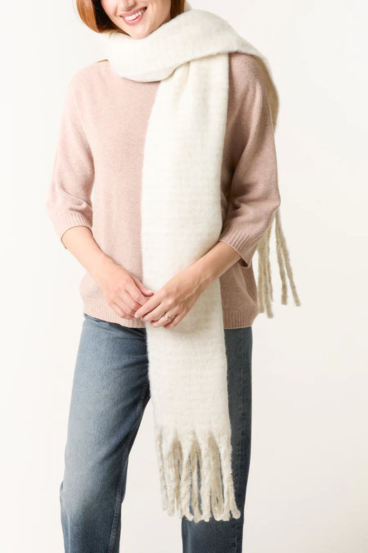 A person wearing a light pink jumper, blue jeans, and a long, thick white vegan tasselled scarf with fringed ends.