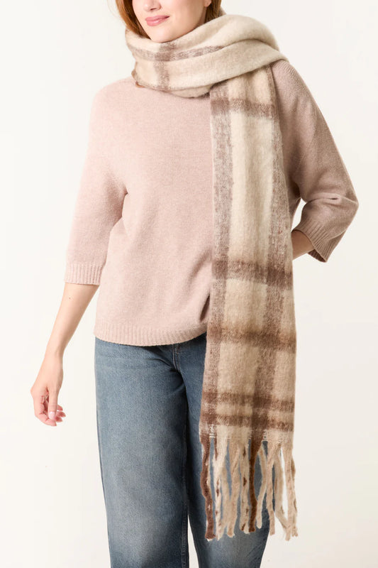 A person wearing a light pink jumper, blue jeans, and a cream checked scarf with fringe draped over their shoulders.