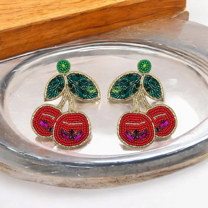 A pair of Beaded Crystal Disco Cherry Earrings, designed as cherries with red fruit, green leaves, and gold edging, make a striking statement piece displayed on a metallic tray.