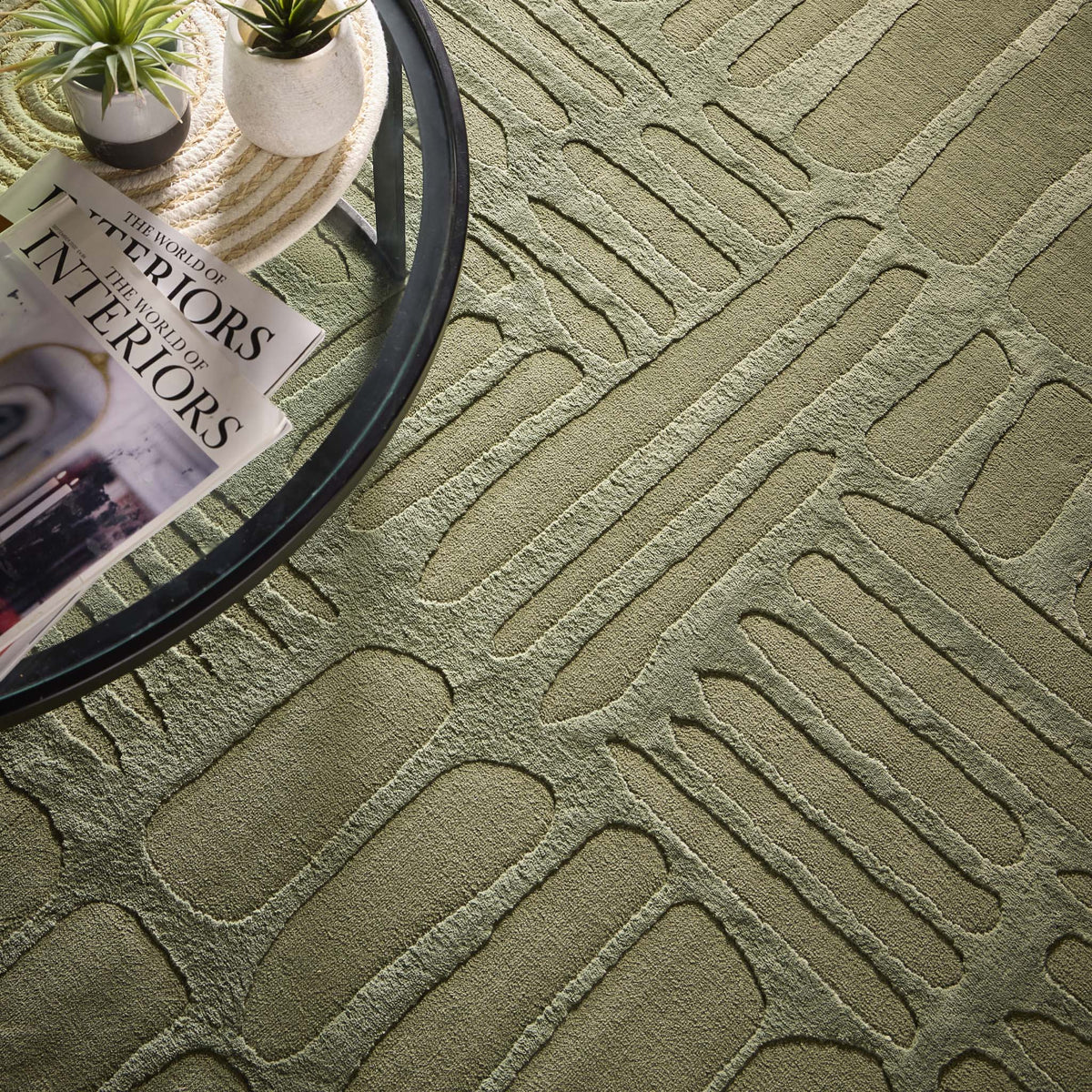 A round glass table with magazines, a small plant, and a modern abstract rug displaying a textured green pattern and non-slip backing beneath.