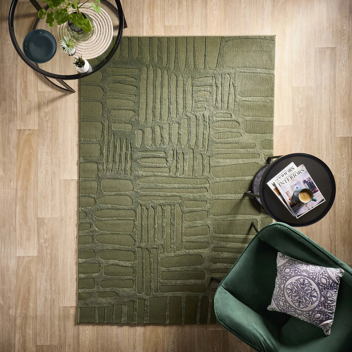 Green modern abstract rug on a wooden floor with a round black side table, a green chair with a patterned cushion, and a plant on another small table.