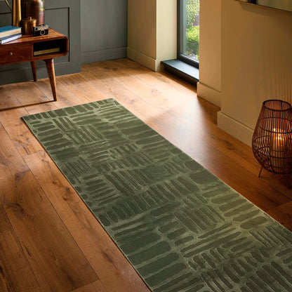 A green patterned modern abstract rug is laid on a wooden floor in a hallway, beside a window, a wooden table, and a decorative lamp.