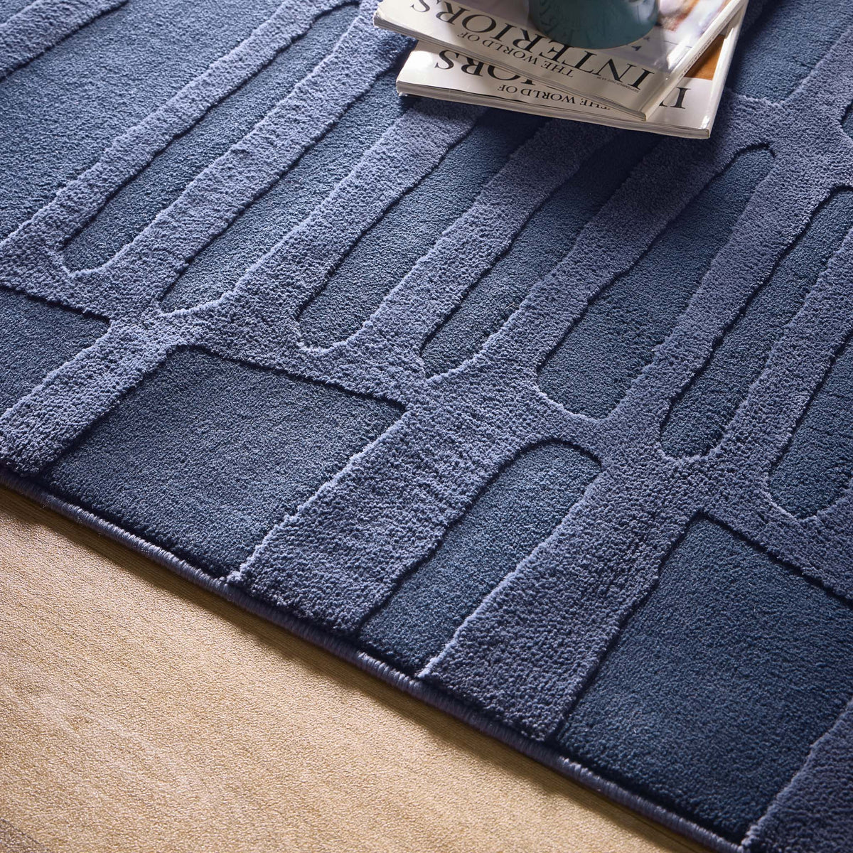 A close-up of a textured dark blue modern abstract rug with a geometric pattern, beside a beige floor. On the non-slip rug lie magazines and a green object, possibly a vase.